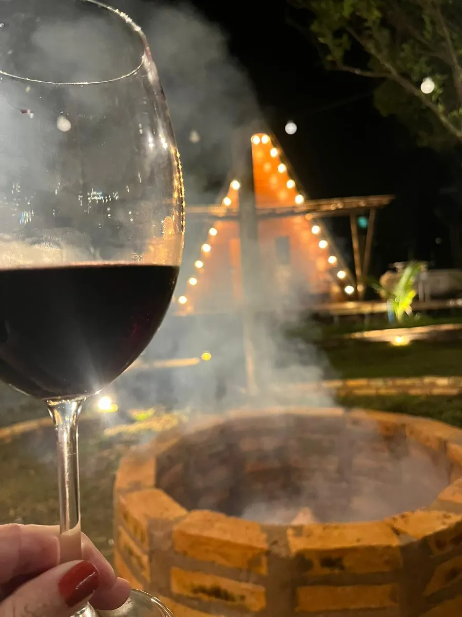 Taça de vinho em frente à fogueira acesa com a cabana iluminada ao fundo